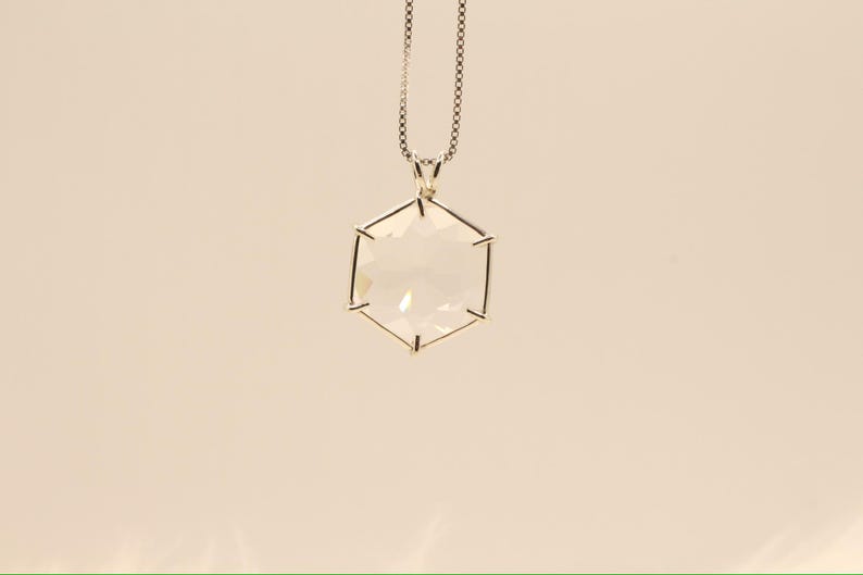 Op de afbeelding: Een zilveren ketting met een heldere, zeshoekige hanger. De hanger hangt aan een fijne ketting. Het geometrische ontwerp en het heldere kristal cre&euml;ren een moderne en elegante uitstraling. De ketting is geschikt voor dagelijks gebruik of speciale gelegenheden.