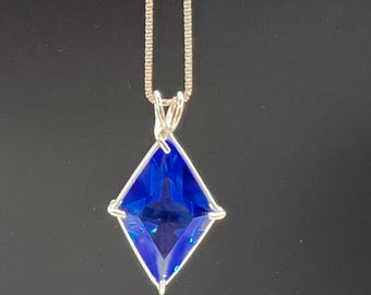 May include: A silver necklace featuring a square, deep blue pendant. The pendant is a faceted gemstone, catching the light. The chain is a delicate silver box chain, and the background is a solid black.