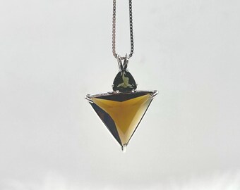 May include: A silver necklace with a geometric pendant. The pendant features a triangular, amber-colored gemstone with a smaller, green gemstone accent. The chain is a delicate, box-style design, and the background is white.