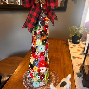 Pode incluir: Decoração artesanal em forma de árvore de Natal. A árvore é coberta por botões coloridos e encimada por um laço xadrez vermelho e preto, raminhos de pinheiro e bagas vermelhas. A base tem formato cilíndrico.
