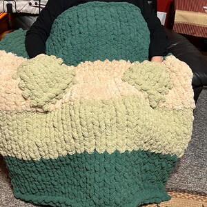May include: A multi-colored chunky knit throw blanket with a green, beige, and pink color scheme. The blanket is draped over a chair and has two armrests made of the same material.