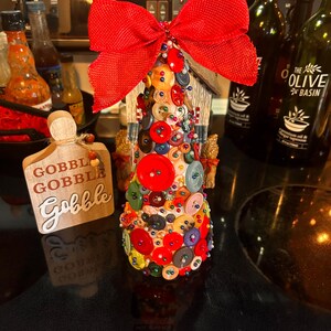 Könnte beinhalten: Ein mit Knöpfen bedeckter Weihnachtsbaum mit einer roten Schleife oben. Der Baum ist mit einer Vielzahl von Knöpfen in verschiedenen Farben und Größen verziert. Der Baum steht auf einer schwarzen Oberfläche mit einem Schild, auf dem "Gobble Gobble Gobble" steht.