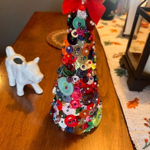 Puede incluir: Un pequeño árbol de Navidad blanco en forma de cono decorado con botones coloridos y cuentas de perlas blancas. Un lazo de terciopelo rojo con un acento dorado se coloca en la parte superior del árbol.