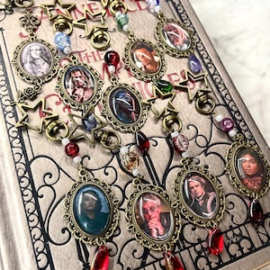 May include: A collection of five antique bronze-toned metal charms featuring oval portrait frames with character images, star-shaped clasps, and dangling red teardrop beads. Each charm is accented with small colorful glass beads and metal links.