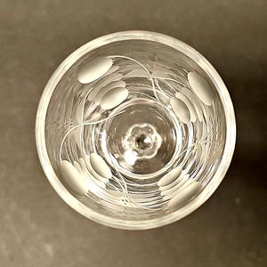 Puede incluir: Una copa de vino de vidrio transparente con un delicado diseño grabado de líneas y círculos en espiral. La copa tiene un tallo corto y una copa ancha.