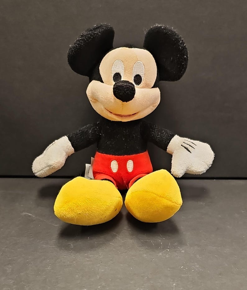 May include: A plush Mickey Mouse toy. The iconic character has a black body, ears, and nose, with a cream-colored face and white gloves. He wears red shorts and yellow shoes. The toy is sitting on a dark surface.