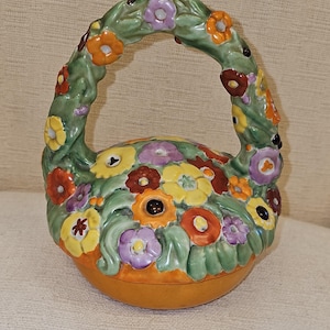 Könnte beinhalten: Eine Keramik-Schmuckschatulle in Form eines Korbes, der mit bunten Blumen überquillt. Der Korb hat einen Griff aus grünen Ranken mit Blumen und Blättern. Die Blumen sind in den Farben Gelb, Orange, Rot, Lila und Grün.