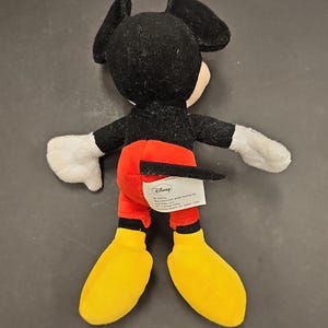 May include: A plush Mickey Mouse toy, viewed from the back. The toy features a black head and ears, a white face, black arms, white gloves, red shorts, yellow shoes, and a black tail. A Disney tag is visible.