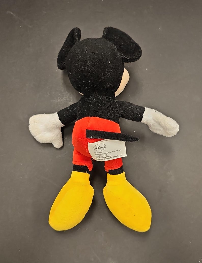 May include: A plush Mickey Mouse toy, viewed from the back. The toy features a black head and ears, a white face, black arms, white gloves, red shorts, yellow shoes, and a black tail. A Disney tag is visible.