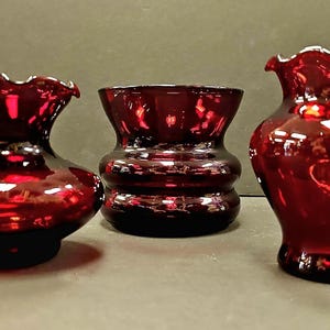 Könnte beinhalten: Drei rubinrote Glasvasen in verschiedenen Größen und Formen. Die Vasen sind alle aus tiefrotem Glas gefertigt und haben eine glänzende Oberfläche.