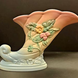 Op de afbeelding: Een keramische hoorn des overvloeds vaas in pastelkleuren. De vaas heeft een gebogen vorm met een uitlopende bovenkant en een voet. Het is versierd met reliëfbloemen in geel en roze, met groene bladeren. De vaas is geschilderd in tinten perzik, geel en blauw.