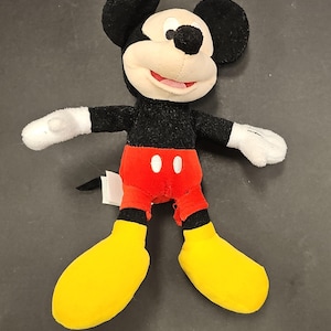 May include: A plush Mickey Mouse doll with black fur, a cream-colored face, and large black ears. He wears red shorts with white buttons, yellow shoes, and white gloves. The doll has a smiling expression and is a classic Disney character.