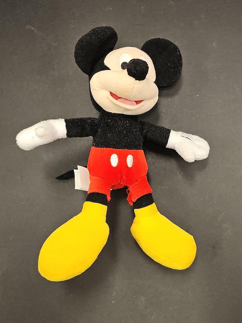 May include: A plush Mickey Mouse doll with black fur, a cream-colored face, and large black ears. He wears red shorts with white buttons, yellow shoes, and white gloves. The doll has a smiling expression and is a classic Disney character.