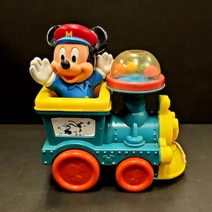 Puede incluir: Un tren de juguete de plástico vintage con una figura de Mickey Mouse en la cabina. El tren es azul, amarillo y rojo con una cúpula de plástico transparente en la parte superior. La cúpula contiene bolas de colores que hacen ruido cuando se mueve el tren.