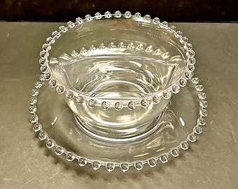 May include: A clear glass divided serving bowl with a beaded rim sits on a matching clear glass plate with a beaded rim.