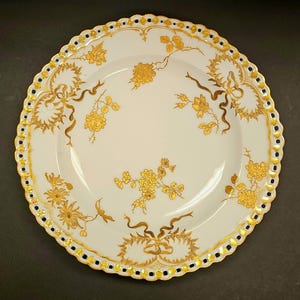 May include: White porcelain plate with a scalloped edge, adorned with gold floral and ribbon designs. The rim features a gold border with black accents. A decorative piece for serving or display.