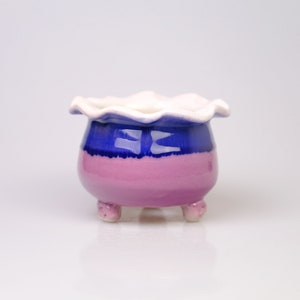 May include: A small, three-legged ceramic planter with a ruffled rim. The planter is glazed in shades of pink, blue, and white.