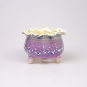 May include: A small, purple ceramic planter with a scalloped rim and three legs. The planter has a white glaze with a speckled pattern.