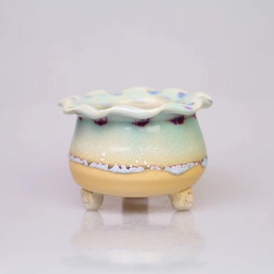 May include: A small, ceramic planter with a scalloped rim and three legs. The planter is decorated with a light blue glaze and white speckles. The glaze has a subtle, iridescent quality.