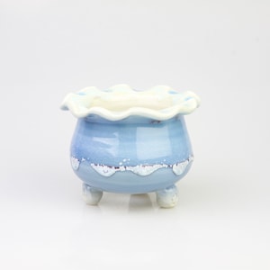 May include: A light blue ceramic planter with a white rim and three small feet. The planter has a scalloped edge and a distressed finish.