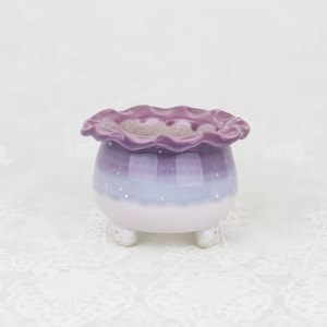 May include: A small ceramic plant pot with a frilled edge, painted in shades of purple, lavender, and white. The pot has three small feet and a speckled design. Suitable for small plants or succulents.
