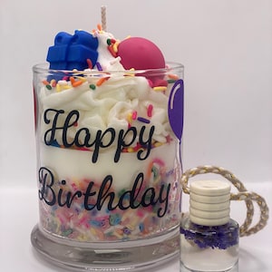 May include: A birthday-themed candle in a clear glass jar, decorated with white frosting, colorful sprinkles, and a blue gift-shaped wax accent. The words "Happy Birthday" are written on the jar. A small glass bottle with a wooden cap and a gold rope is next to the candle.