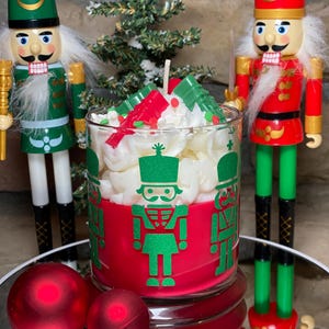 May include: A festive candle in a glass jar with a red base and green nutcracker soldier graphics. The candle is topped with white wax and red and green decorative accents. Two nutcracker figurines and red ornaments are in the background.