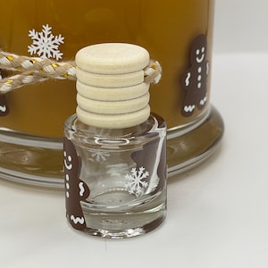 May include: A small clear glass bottle with a light-colored wooden cap and a braided cord, featuring brown gingerbread man decals and white snowflake graphics. Behind it sits a larger brown container with similar gingerbread and snowflake designs.