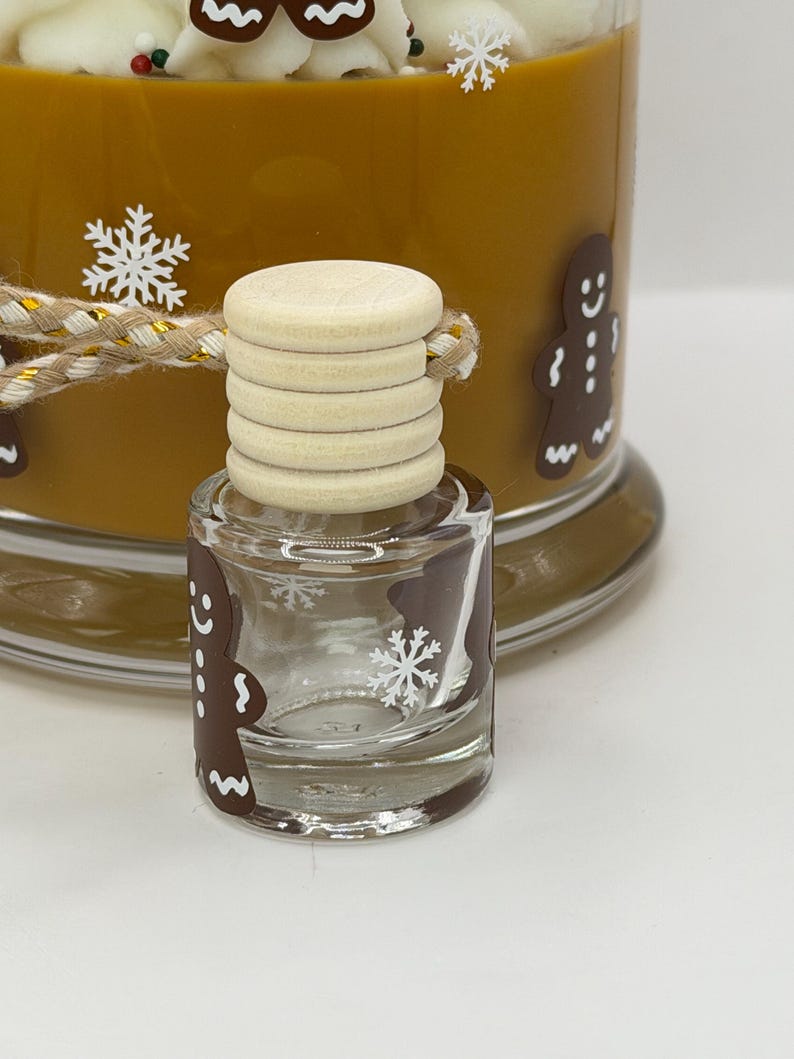 May include: A small clear glass bottle with a light-colored wooden cap and a braided cord, featuring brown gingerbread man decals and white snowflake graphics. Behind it sits a larger brown container with similar gingerbread and snowflake designs.