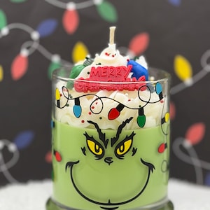 May include: A green candle in a glass jar featuring a Grinch face graphic and a string of colorful holiday lights. The top is decorated with white wax resembling whipped cream, small red sprinkles, and a red sign that reads Merry Grinchmas.