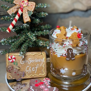 May include: A glass candle container filled with brown wax, topped with white whipped wax, gingerbread man figures, and red and green sprinkles. The glass features white snowflake and candy cane decals. A sign reads Gingerbread Chef.