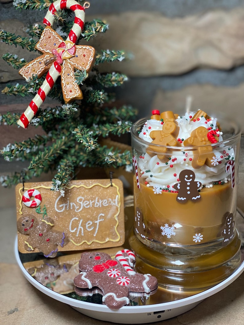 May include: A glass candle container filled with brown wax, topped with white whipped wax, gingerbread man figures, and red and green sprinkles. The glass features white snowflake and candy cane decals. A sign reads Gingerbread Chef.