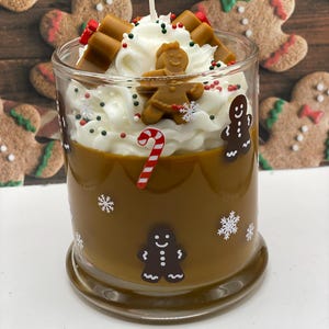 May include: A glass candle filled with brown wax, decorated with gingerbread man decals and white snowflakes. The top features white whipped wax, a small gingerbread figure, candy cane accents, and colorful sprinkles with a central wick.