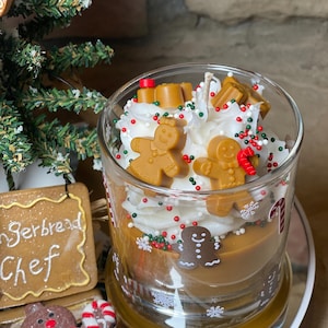 May include: A glass candle filled with brown wax, topped with white whipped wax resembling frosting, decorated with small brown gingerbread person figures, red and green sprinkles, and white snowflake decals on the exterior glass surface.
