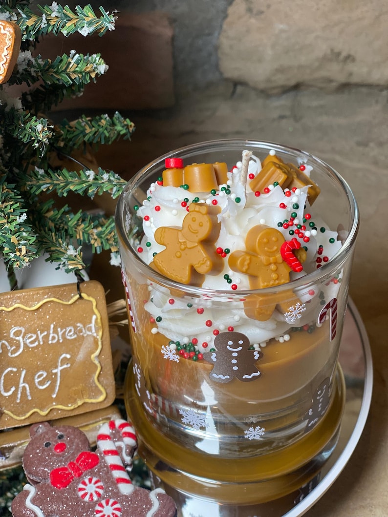 May include: A glass candle filled with brown wax, topped with white whipped wax resembling frosting, decorated with small brown gingerbread person figures, red and green sprinkles, and white snowflake decals on the exterior glass surface.