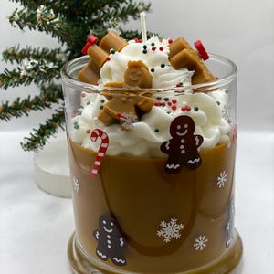 May include: A glass candle jar filled with brown wax, decorated with gingerbread man and snowflake decals. The top features white whipped wax, topped with small gingerbread man figures, red and green sprinkles, and a central wick.