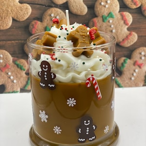 May include: A brown candle in a glass jar decorated with white snowflake decals and gingerbread man figures. The top features white wax piped to resemble whipped cream, topped with small gingerbread man wax embeds, red and green sprinkles, and a candy cane.