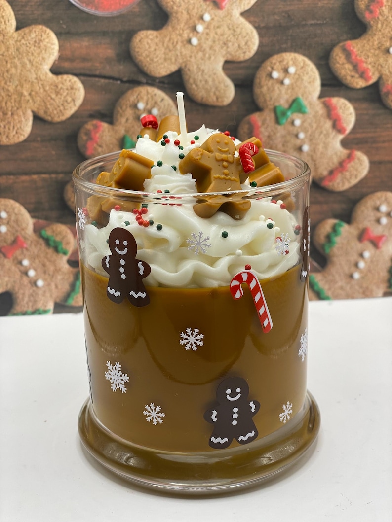 May include: A brown candle in a glass jar decorated with white snowflake decals and gingerbread man figures. The top features white wax piped to resemble whipped cream, topped with small gingerbread man wax embeds, red and green sprinkles, and a candy cane.