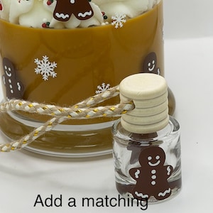 May include: A brown candle with white whipped wax topping, gingerbread man decals, and white snowflakes. A small glass car diffuser with a wooden cap and a gingerbread man decal sits in front, tied with a gold and tan cord. Text reads: Add a matching gingerbread car diffuser For 5.00