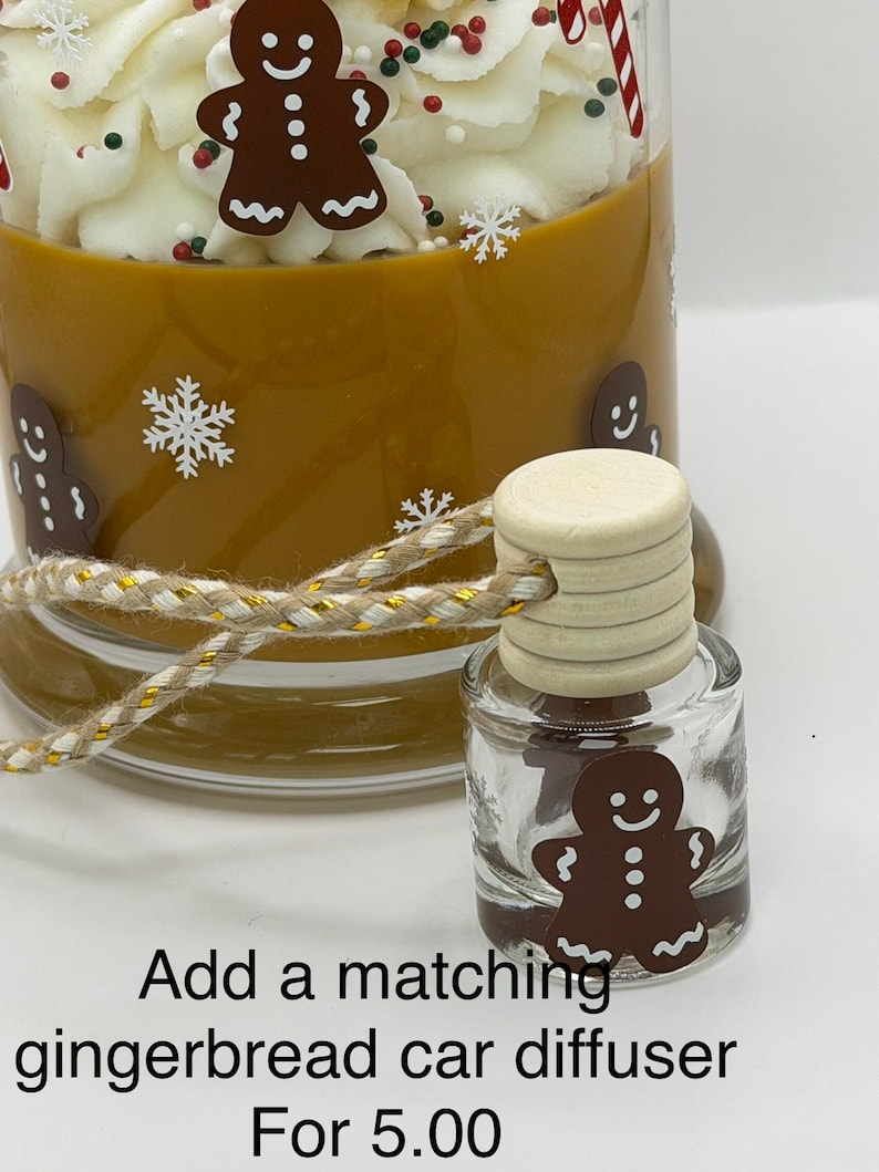 May include: A brown candle with white whipped wax topping, gingerbread man decals, and white snowflakes. A small glass car diffuser with a wooden cap and a gingerbread man decal sits in front, tied with a gold and tan cord. Text reads: Add a matching gingerbread car diffuser For 5.00