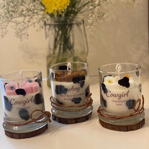May include: Three glass candles with a cowgirl theme. Each candle has a different scent, with the words "Cowgirl" and "Cowboy" printed on the glass. The candles are decorated with black and white spots and sit on a wooden base.