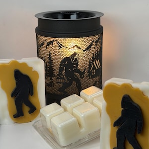 May include: A black metal wax warmer with a cutout silhouette of a Bigfoot figure holding a beer, mountains, and trees. Two rectangular wax melts feature a yellow Bigfoot shape on a white background, and a clear plastic tray holds six white wax cubes.