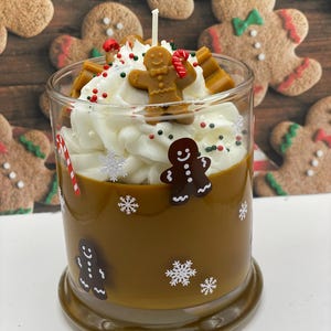 May include: A brown candle in a glass jar decorated with gingerbread man and snowflake decals. The top features white wax piped to resemble whipped cream, topped with a small gingerbread man figure, candy cane pieces, and sprinkles.