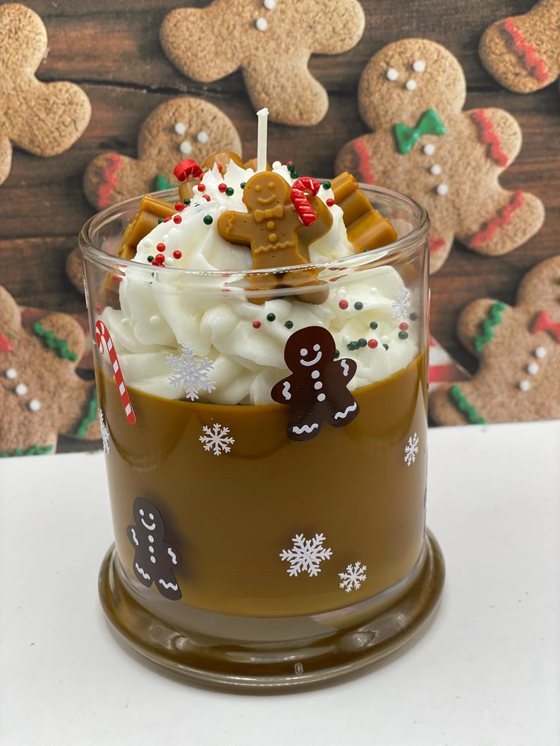May include: A brown candle in a glass jar decorated with gingerbread man and snowflake decals. The top features white wax piped to resemble whipped cream, topped with a small gingerbread man figure, candy cane pieces, and sprinkles.