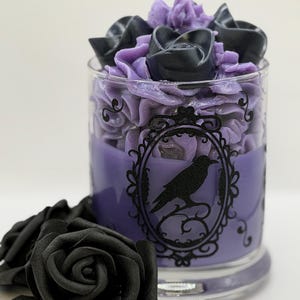 May include: A glass candle container filled with purple wax, topped with piped purple and black wax roses and a central wick. The glass features a black printed oval frame containing a silhouette of a raven. Two black artificial roses sit nearby.