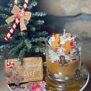 May include: A glass candle container filled with tan wax, topped with white whipped wax, small gingerbread man figures, and colorful sprinkles. The glass features gingerbread man and snowflake decals. A gingerbread-shaped sign reads Gingerbread Chef.