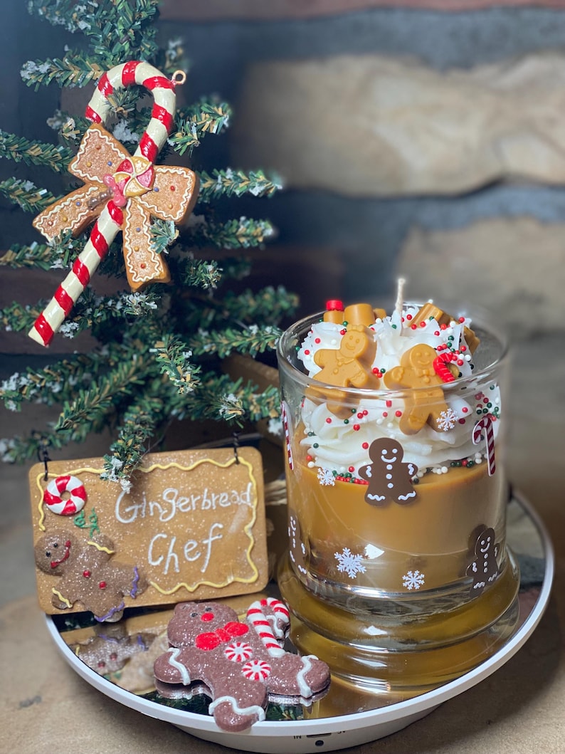 May include: A glass candle container filled with tan wax, topped with white whipped wax, small gingerbread man figures, and colorful sprinkles. The glass features gingerbread man and snowflake decals. A gingerbread-shaped sign reads Gingerbread Chef.