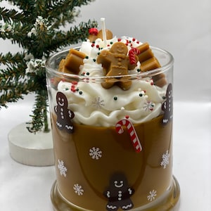 May include: A glass candle container filled with brown wax, decorated with gingerbread man decals and white snowflakes. The top features white whipped wax, a central wick, gingerbread man wax embeds, and red and green sprinkles.