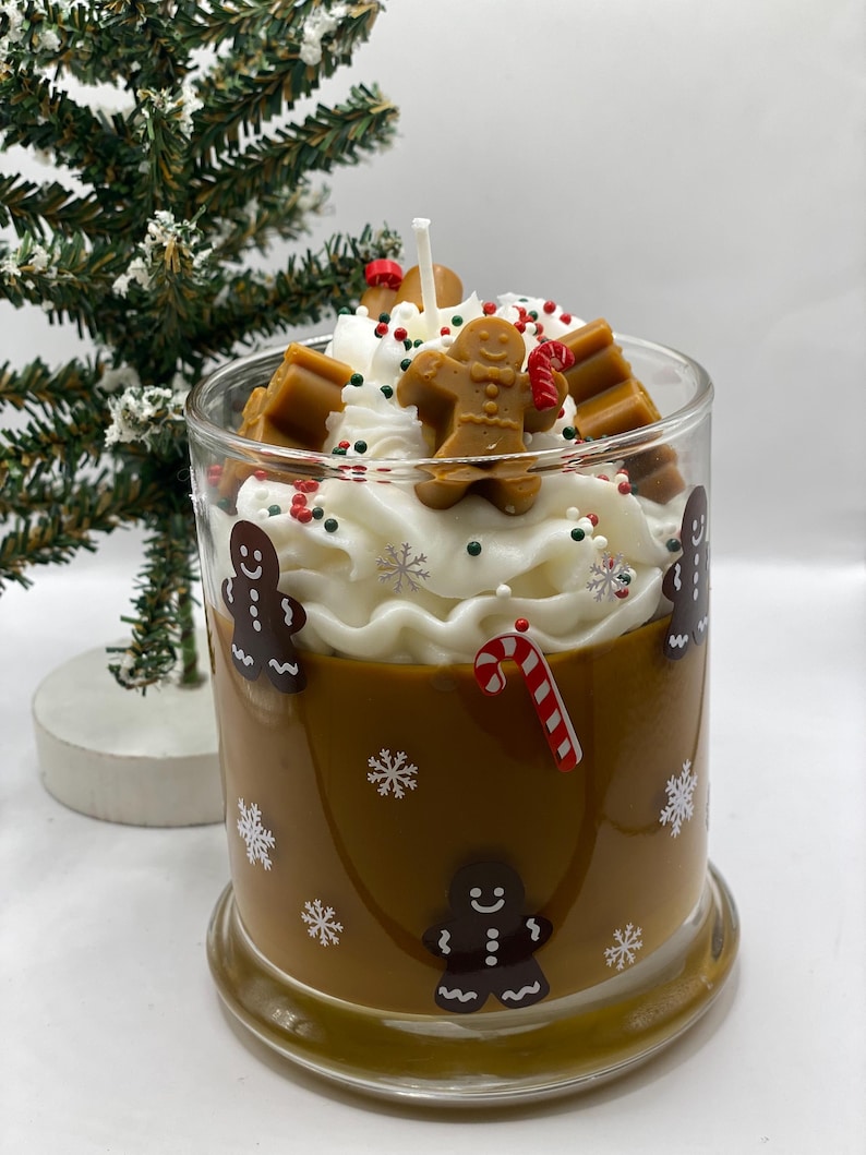 May include: A glass candle container filled with brown wax, decorated with gingerbread man decals and white snowflakes. The top features white whipped wax, a central wick, gingerbread man wax embeds, and red and green sprinkles.