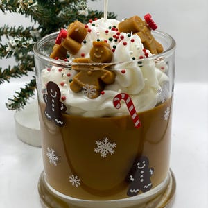 May include: A glass candle container filled with brown wax, decorated with white snowflake decals and gingerbread man graphics. The top features white whipped wax, brown gingerbread man wax embeds, red and green sprinkles, and a candy cane.